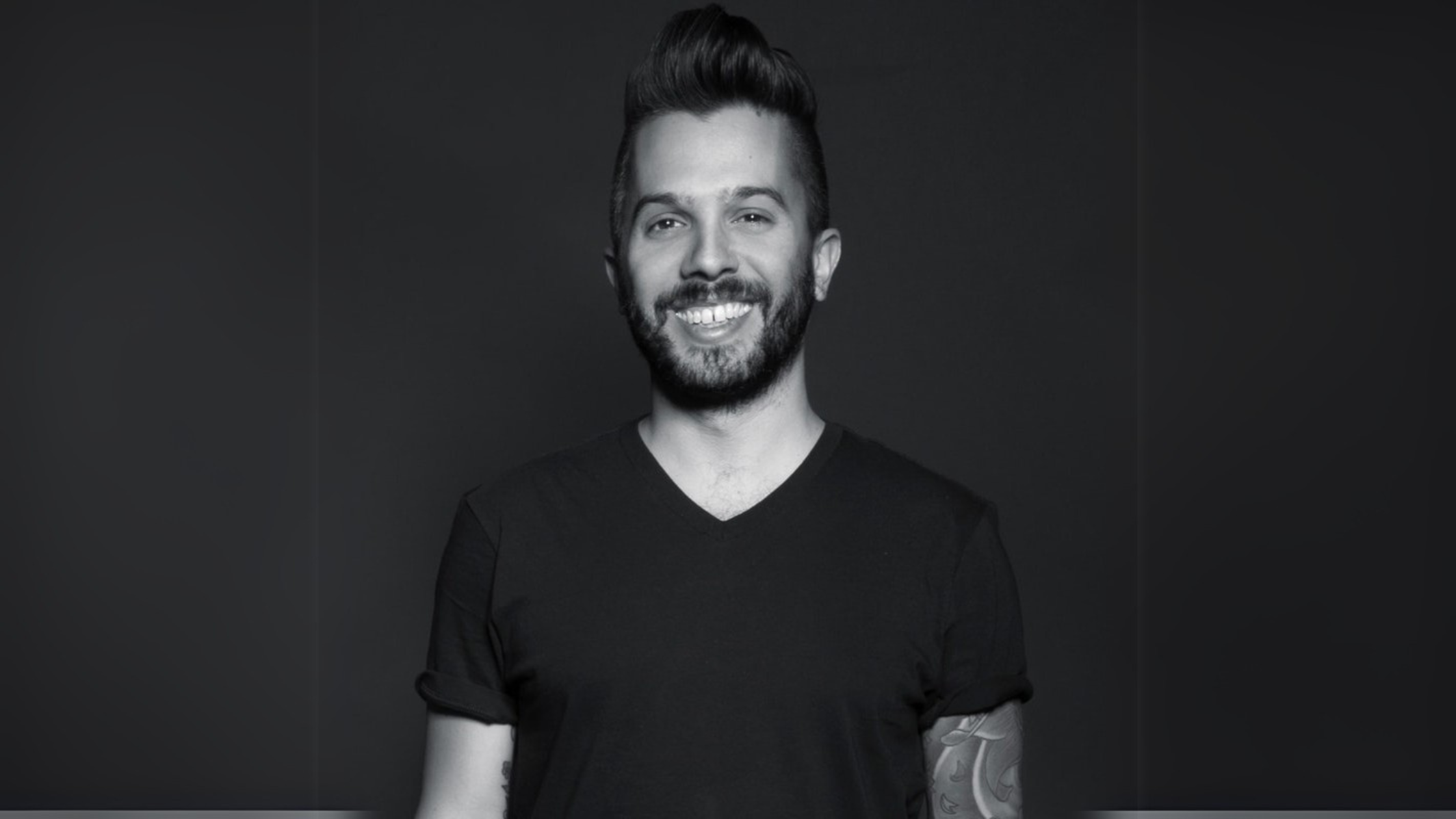 Black and white portrait of John Sampogna, Co-Founder & CEO of Wondersauce, smiling while wearing a black V-neck T-shirt. His tattooed arm is partially visible, and the background is dark and minimalistic.