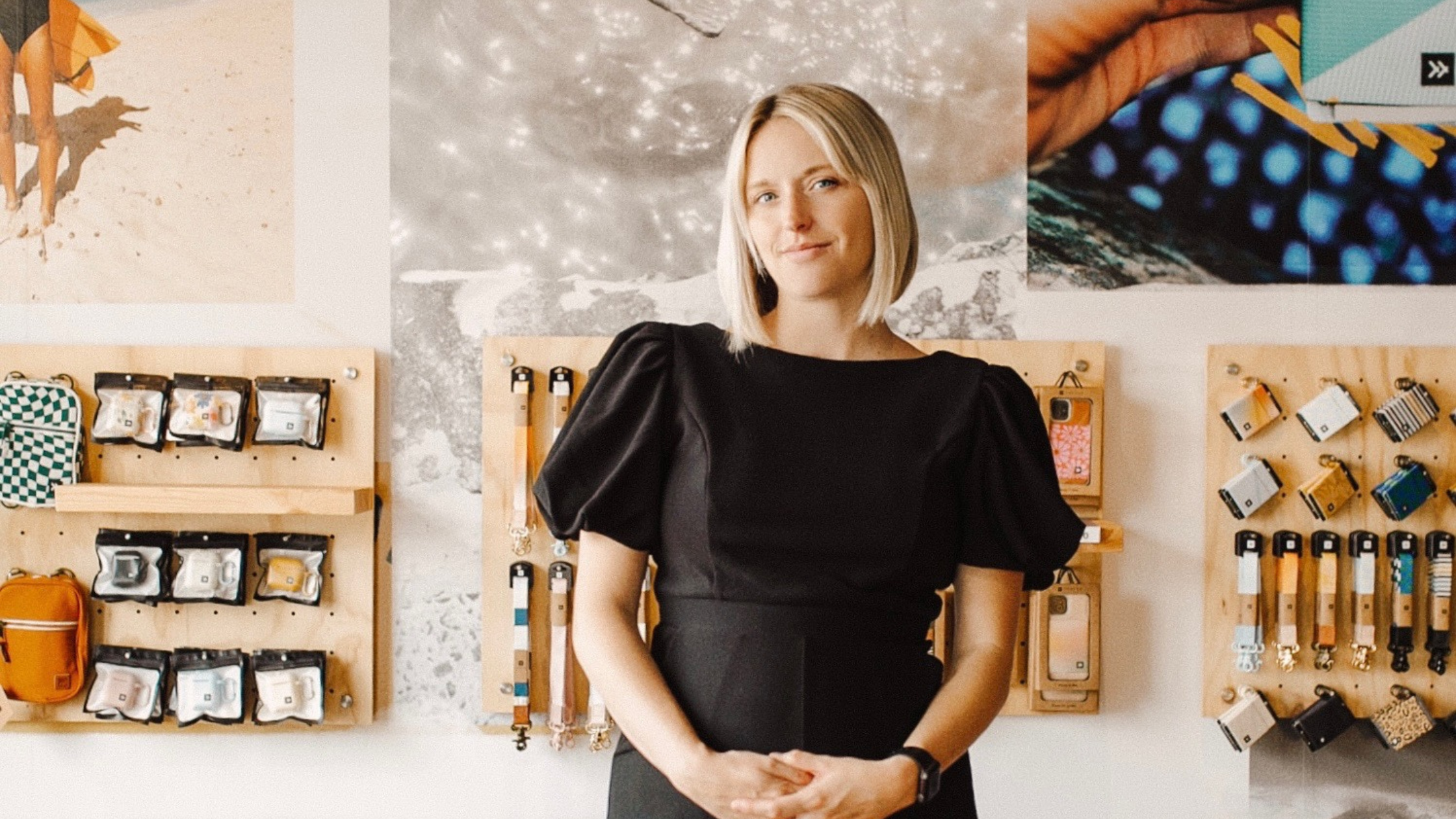 A portrait of McKenzie King Bauer, Co-Founder of Thread, standing confidently in front of a display of her company's carry accessories. She wears a black dress with puffed sleeves, and her blonde bob frames her face. The background features wooden pegboard displays showcasing wallets, keychains, and small bags, along with large lifestyle images that reflect Thread’s brand aesthetic. The setting conveys a modern and stylish retail or showroom space.