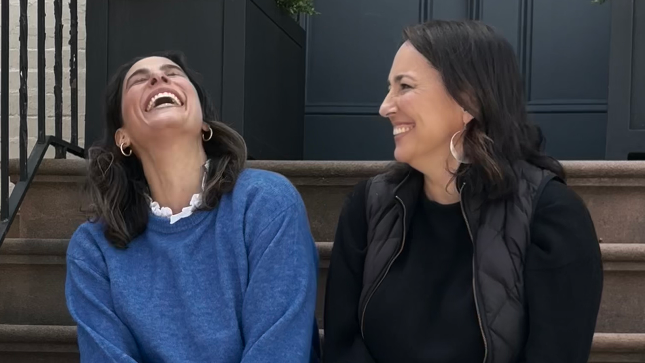 Two women sit on brownstone steps, sharing a joyful moment. The woman on the left, wearing a blue sweater with a white ruffled collar and gold hoop earrings, throws her head back in laughter. The woman on the right, dressed in black with large white earrings and a black quilted vest, smiles warmly at her. A dark blue door and railing are visible in the background.
