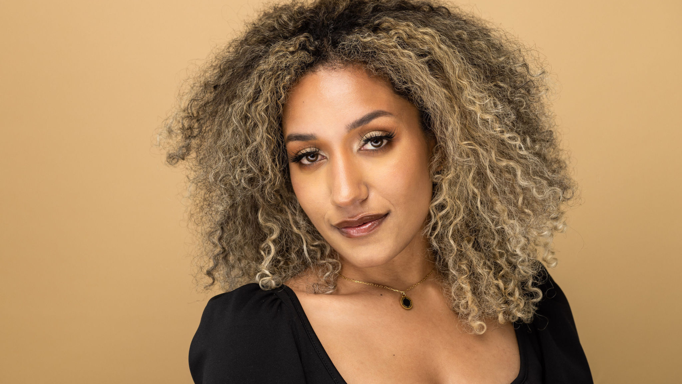 A confident woman, Ila Corcoran, with voluminous, highlighted curly hair and flawless makeup gazes calmly at the camera against a solid tan background. She wears a black top with a square neckline and a delicate gold necklace featuring a black pendant. The portrait conveys a sense of elegance and strength.