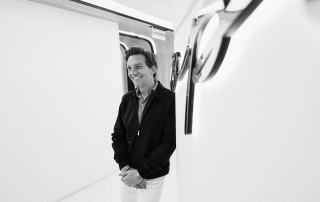 Black-and-white photograph of Dr. Michael Apa standing in a sleek, modern, white hallway with curved walls and contemporary light fixtures. He is smiling, wearing a dark jacket over a collared shirt with light-colored pants, and has his hands clasped loosely in front of him.