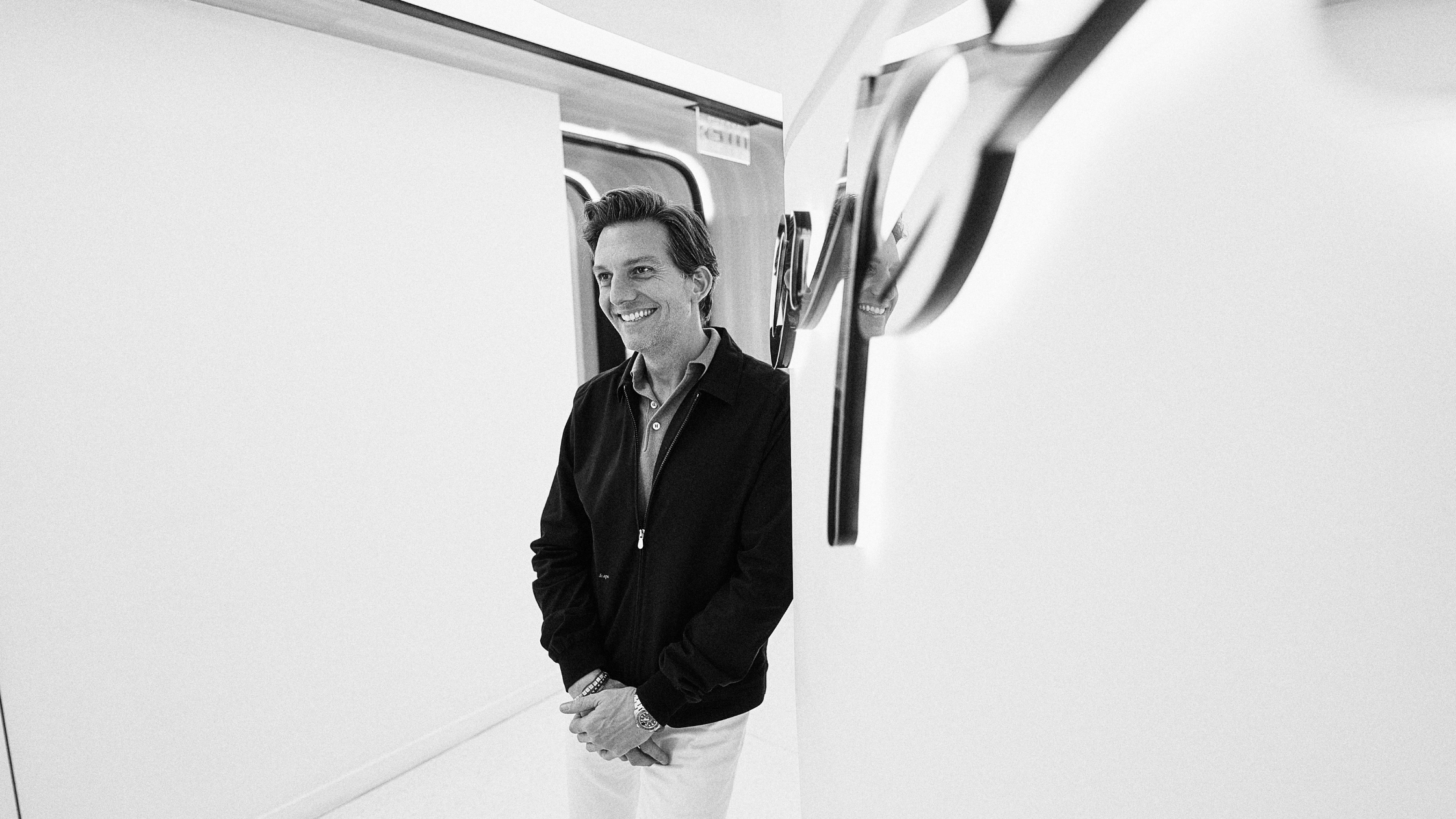 Black-and-white photograph of Dr. Michael Apa standing in a sleek, modern, white hallway with curved walls and contemporary light fixtures. He is smiling, wearing a dark jacket over a collared shirt with light-colored pants, and has his hands clasped loosely in front of him.