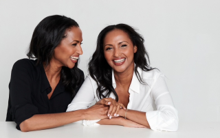 Portrait of twin sisters Feven and Helena Yohannes seated at a white table, smiling and holding hands. One wears a black blouse and the other a white blouse, sharing a moment of laughter and connection.
