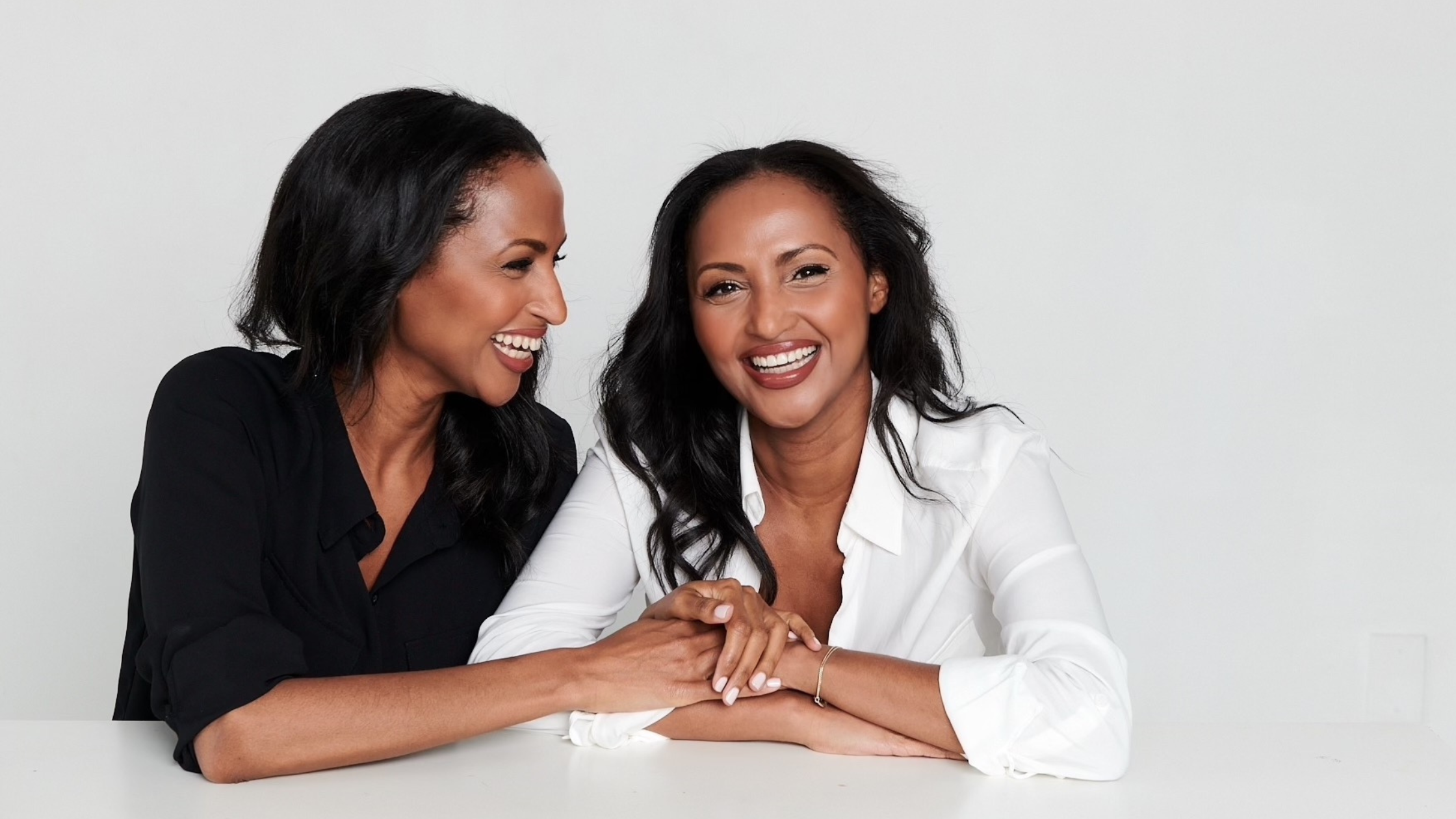 Portrait of twin sisters Feven and Helena Yohannes seated at a white table, smiling and holding hands. One wears a black blouse and the other a white blouse, sharing a moment of laughter and connection.